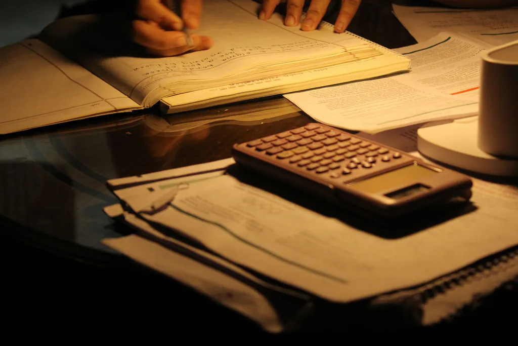 A calculator and stacks of paperwork on a table under warm lamplight late at night, while hands write figures
